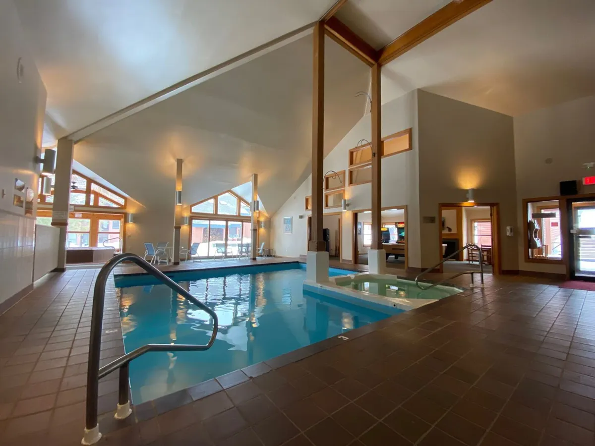 The interior features an indoor swimming pool with a jacuzzi, large windows letting in natural light, and rustic wooden beams. The mood is tranquil and refreshing, ideal for relaxing after outdoor activities.