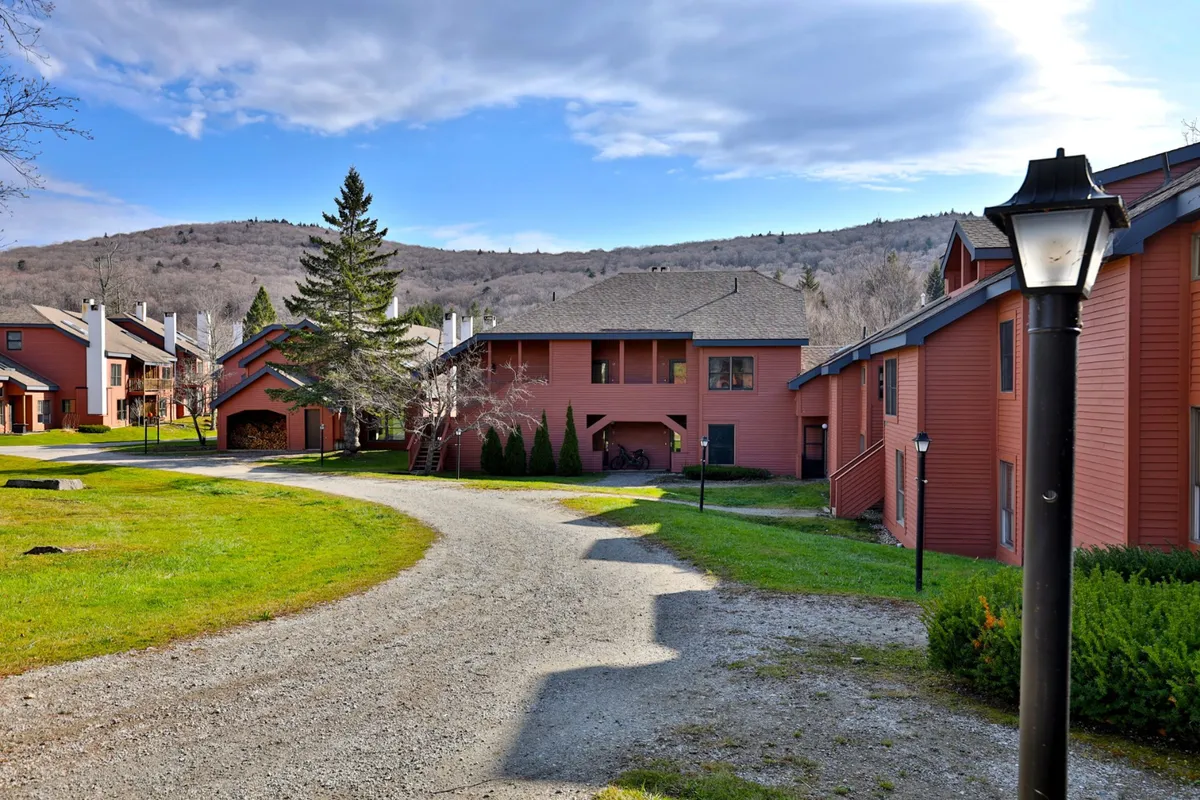 This image features the exterior of red buildings set against a mountain backdrop with a scenic pathway and lamp post, surrounded by green grass. The environment feels inviting and peaceful, ideal for outdoor activities like walking, biking, and enjoying nature.