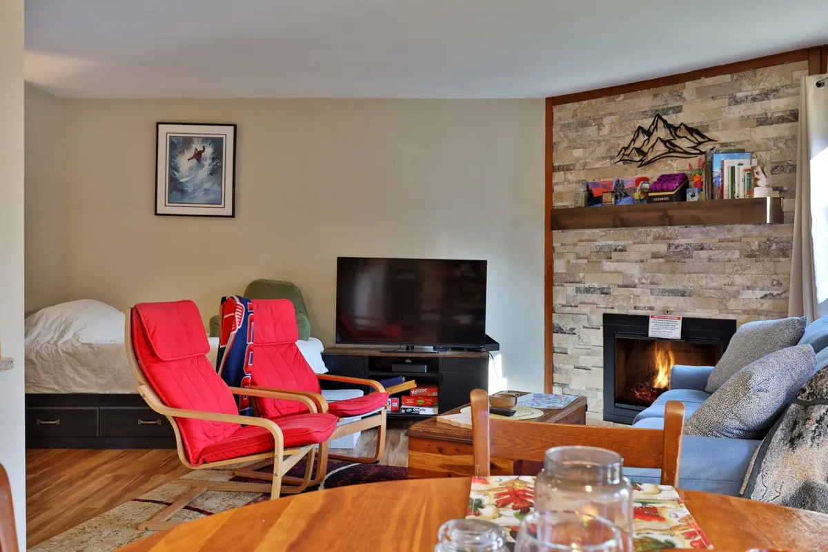 This inviting living room features two plush red chairs positioned near a stone fireplace, a wall-mounted television, and a bookshelf filled with books and decor. The warm firelight and comfortable seating create a relaxed, communal atmosphere ideal for unwinding after outdoor adventures.