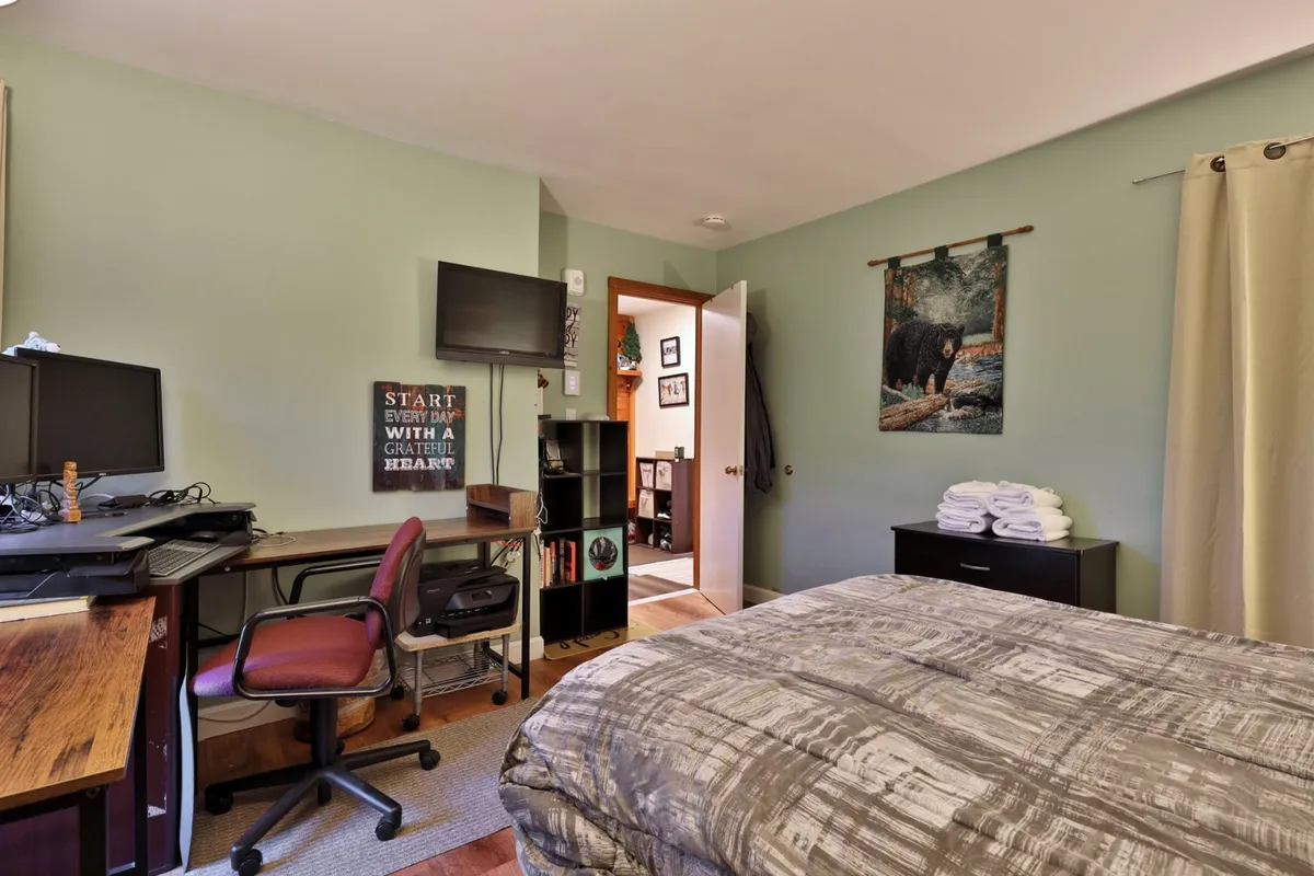 This image showcases a bed with a patterned comforter, a desk with a computer setup, and a bear-themed wall hanging. The room feels inviting and restful, emphasizing comfort and functionality.