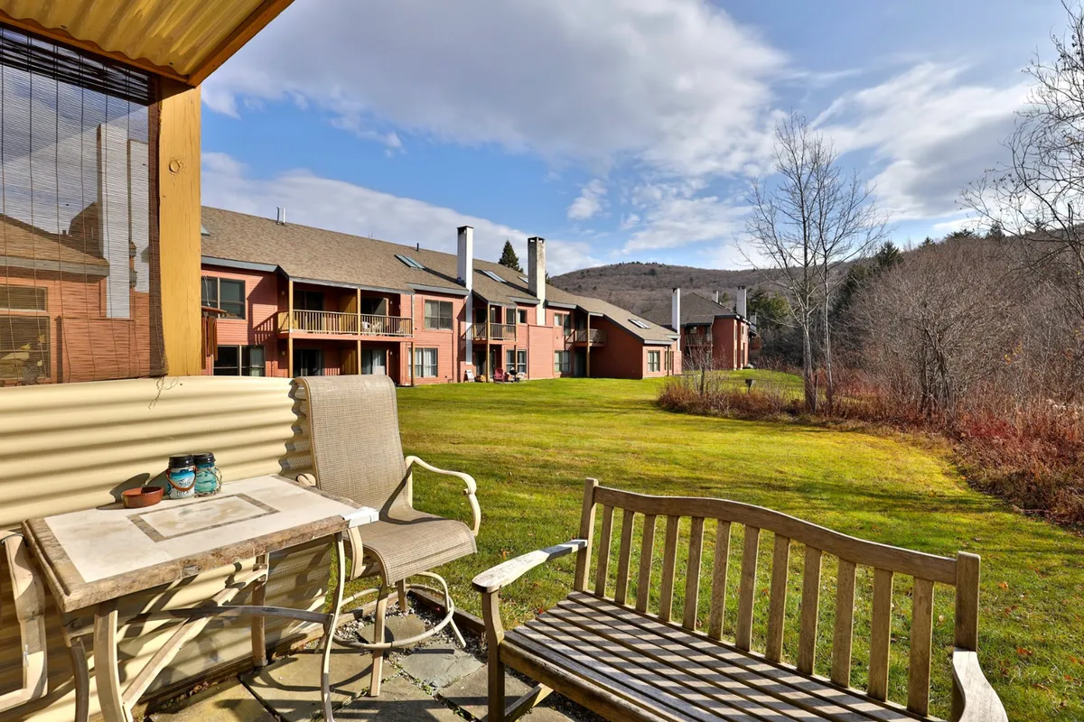 A serene private patio with a table and bench, set on a green lawn against a backdrop of distant mountains and the building's facade. The space feels secluded and ideal for quiet moments or casual gatherings.