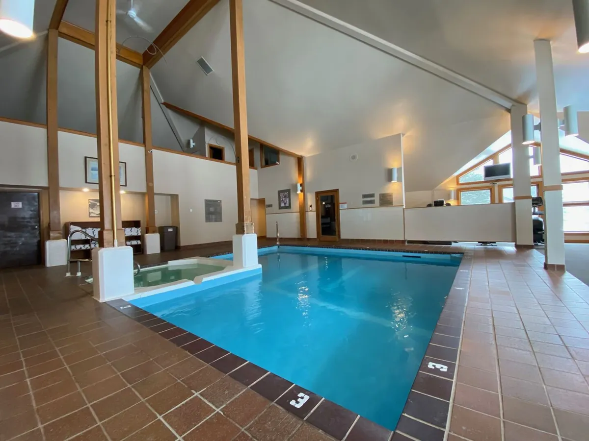 A tranquil indoor space featuring a spacious swimming pool surrounded by tall wooden beams, comfortable seating, and a reception area. Natural light fills the area, creating a welcoming and rejuvenating atmosphere.