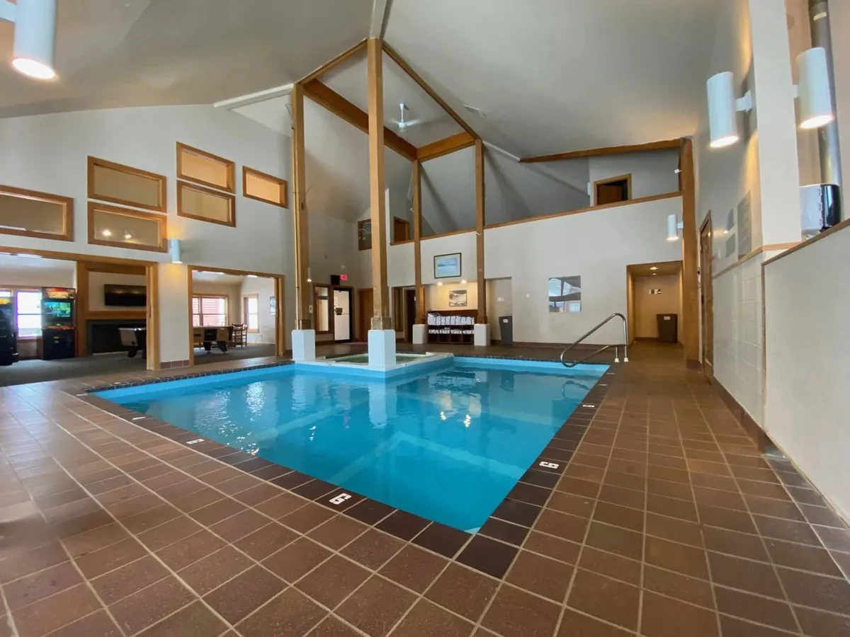 The image shows a spacious indoor pool area with large windows letting in natural light, wooden beams for a rustic touch, and a comfortable seating area. The pool is inviting and there is ample space for guests to relax by the water or socialize in small groups.