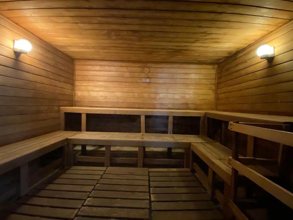 A serene sauna room with wooden benches and walls, softly lit with light fixtures that create a calming atmosphere. The space is designed for relaxation and conversation, embodying wellness and tranquility.