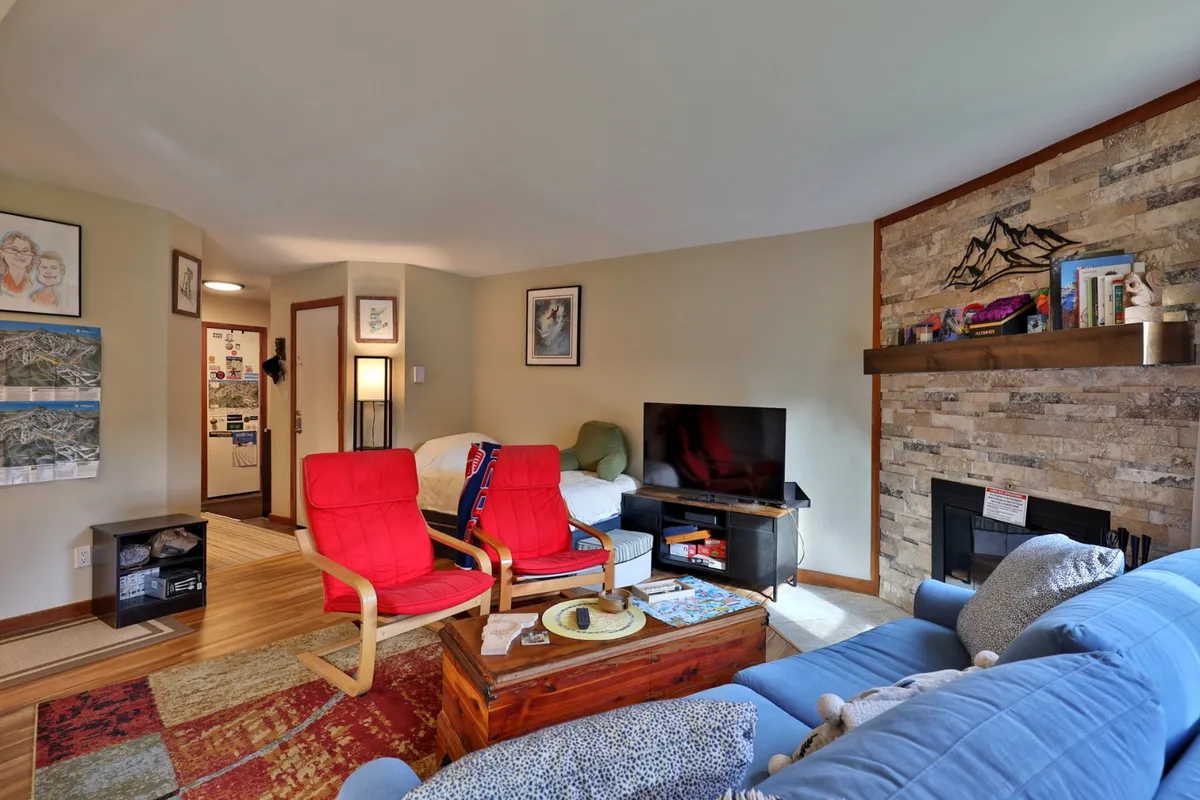 A cozy lounge area featuring red chairs, a stone fireplace, a blue couch, and a wood coffee table. The room invites relaxation, socializing, or enjoying a quiet moment by the fire, with a casual yet warm atmosphere.